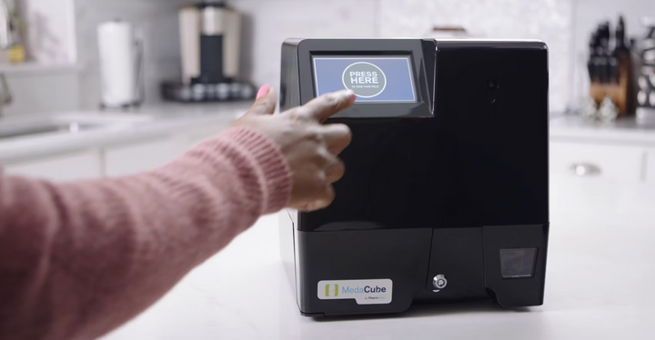 Shows a finger pressing the touch screen on the MedaCube. The screen shows a press here button showing that medications are ready to take. 