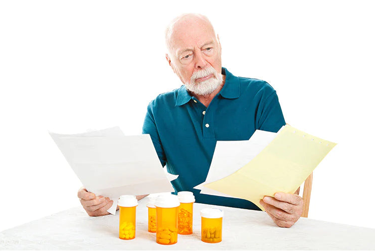 An older man with a white beard sits at a table reading prescription papers, with several orange prescription pill bottles in front of him on a white background.