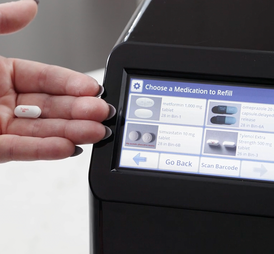 Hand holding a pill next to a medication refilling device with a screen displaying refill options.